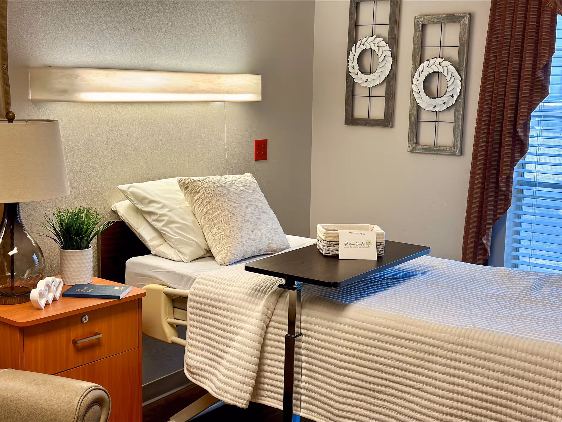 Patient room with a made hospital-style bed, overbed table, bedside lamp, nightstand and wall decorations by a window.