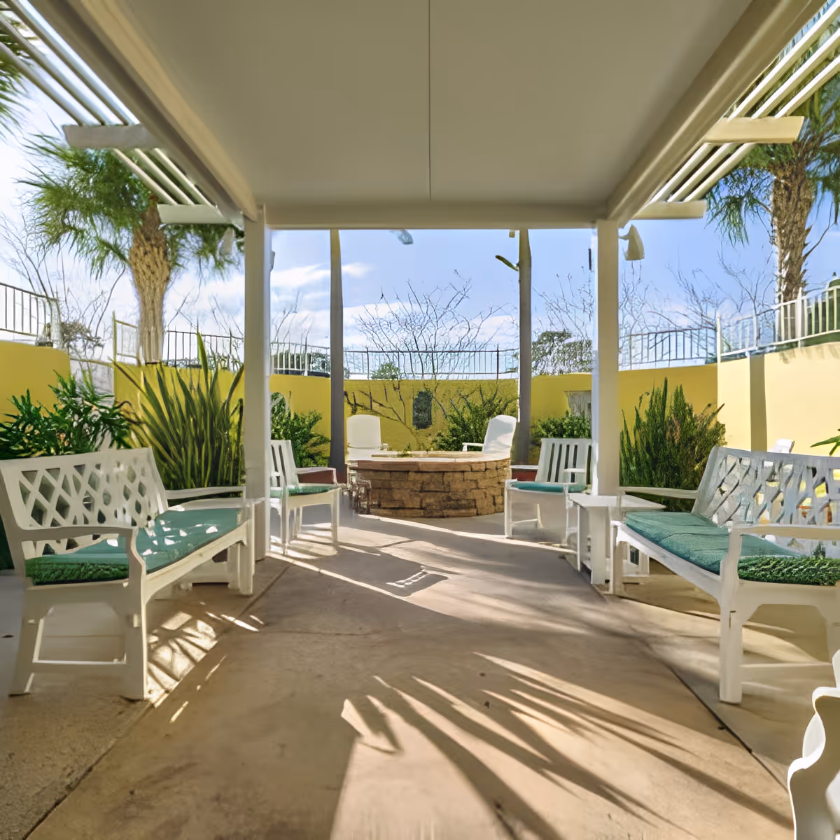 Outdoor covered seating area with white benches and chairs featuring green cushions, surrounding a circular stone fire pit. The area is enclosed by a yellow wall with plants and trees visible beyond it under a clear blue sky.