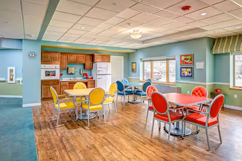 A bright and colorful dining area in The Gardens Retirement Community featuring multiple tables with chairs in yellow, red, and blue. The room has wooden flooring, light blue and green walls, large windows letting in natural light, and a kitchen area with wooden cabinets, a white refrigerator, and a microwave.