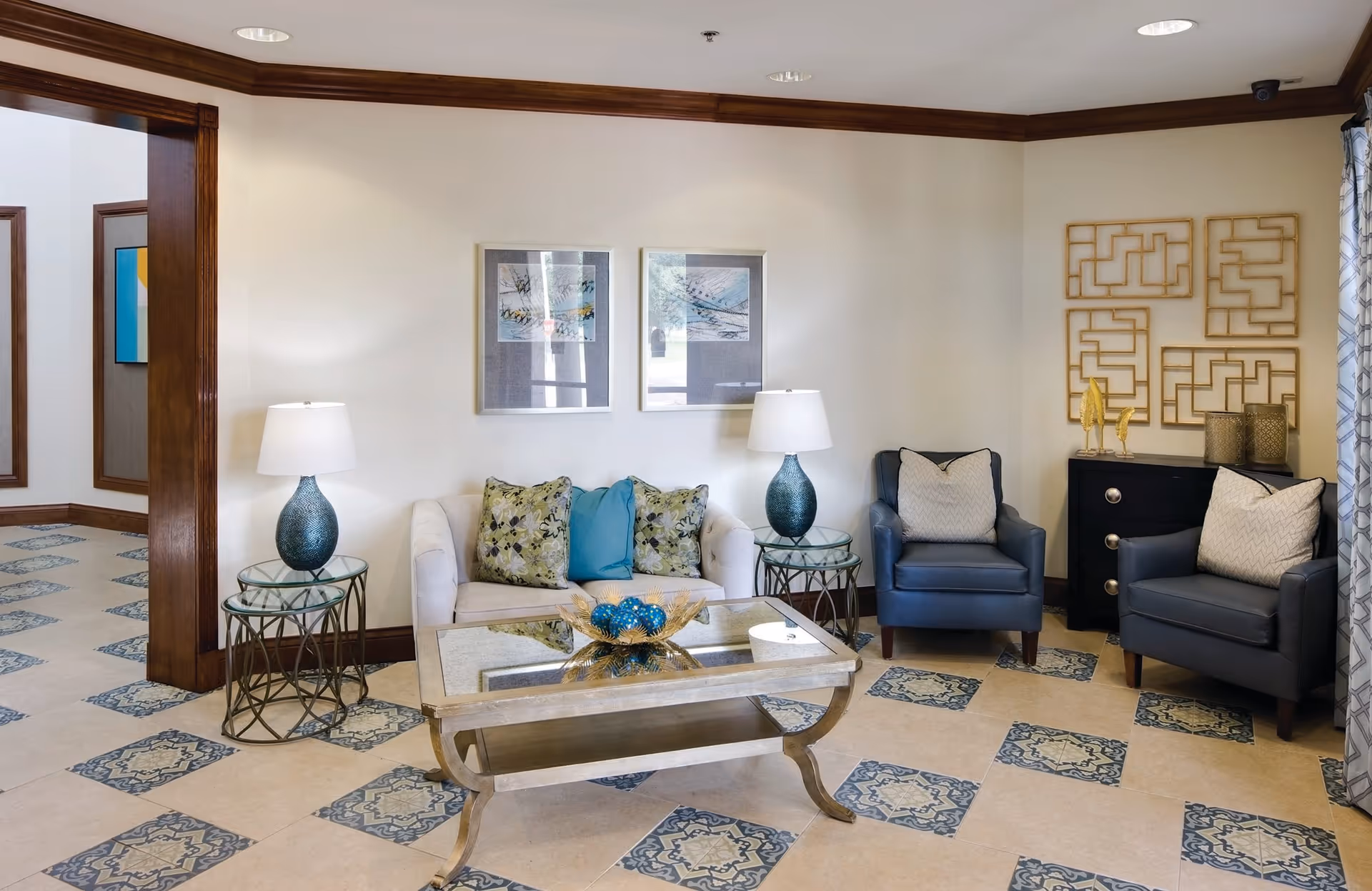 A cozy living room area with a white sofa adorned with patterned and solid blue pillows, two blue armchairs with light-colored cushions, a glass coffee table with decorative blue spheres, two side tables each with a blue lamp, and wall art including framed pictures and geometric gold designs. The room has tiled flooring with blue patterned accents and wood trim around the ceiling and doorways.