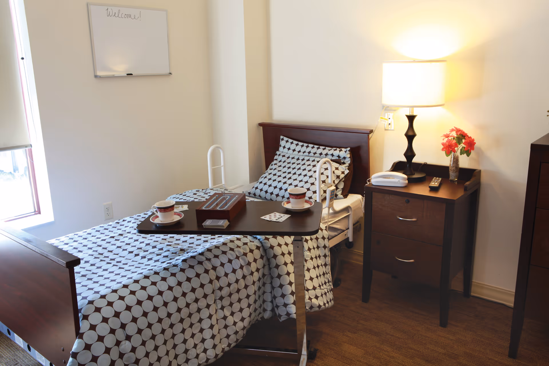 Neat senior bedroom with an adjustable bed, overbed table holding teacups, and a bedside table with a lamp and flowers.