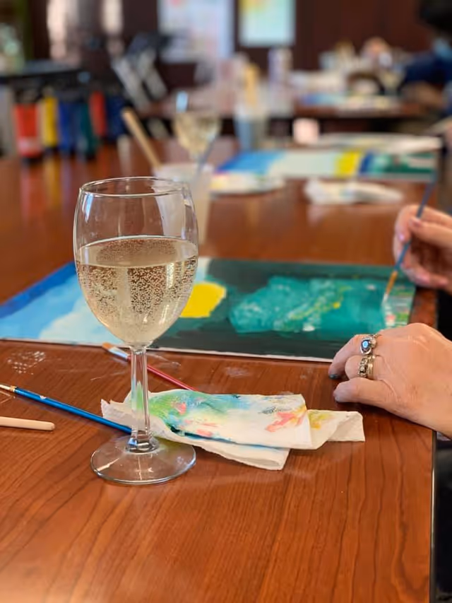 Close-up of a glass of white wine on a wooden table with paintbrushes and a paint-stained napkin nearby. In the background, a person is painting on a canvas with green and yellow colors.