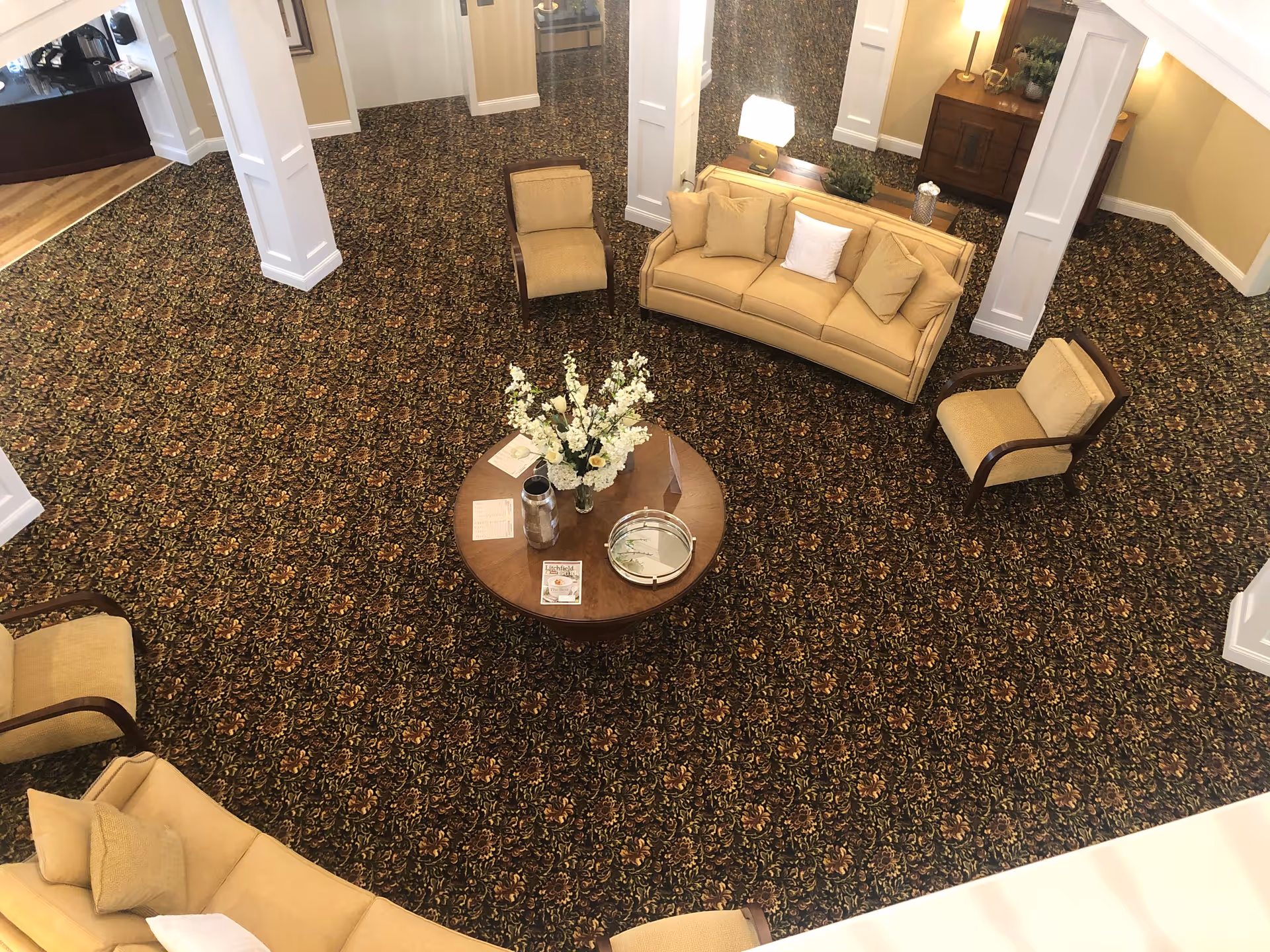 Overhead view of a living room area with patterned carpet, beige sofas and armchairs arranged around a round wooden coffee table with a flower arrangement and decorative items. The room has white pillars and warm lighting.