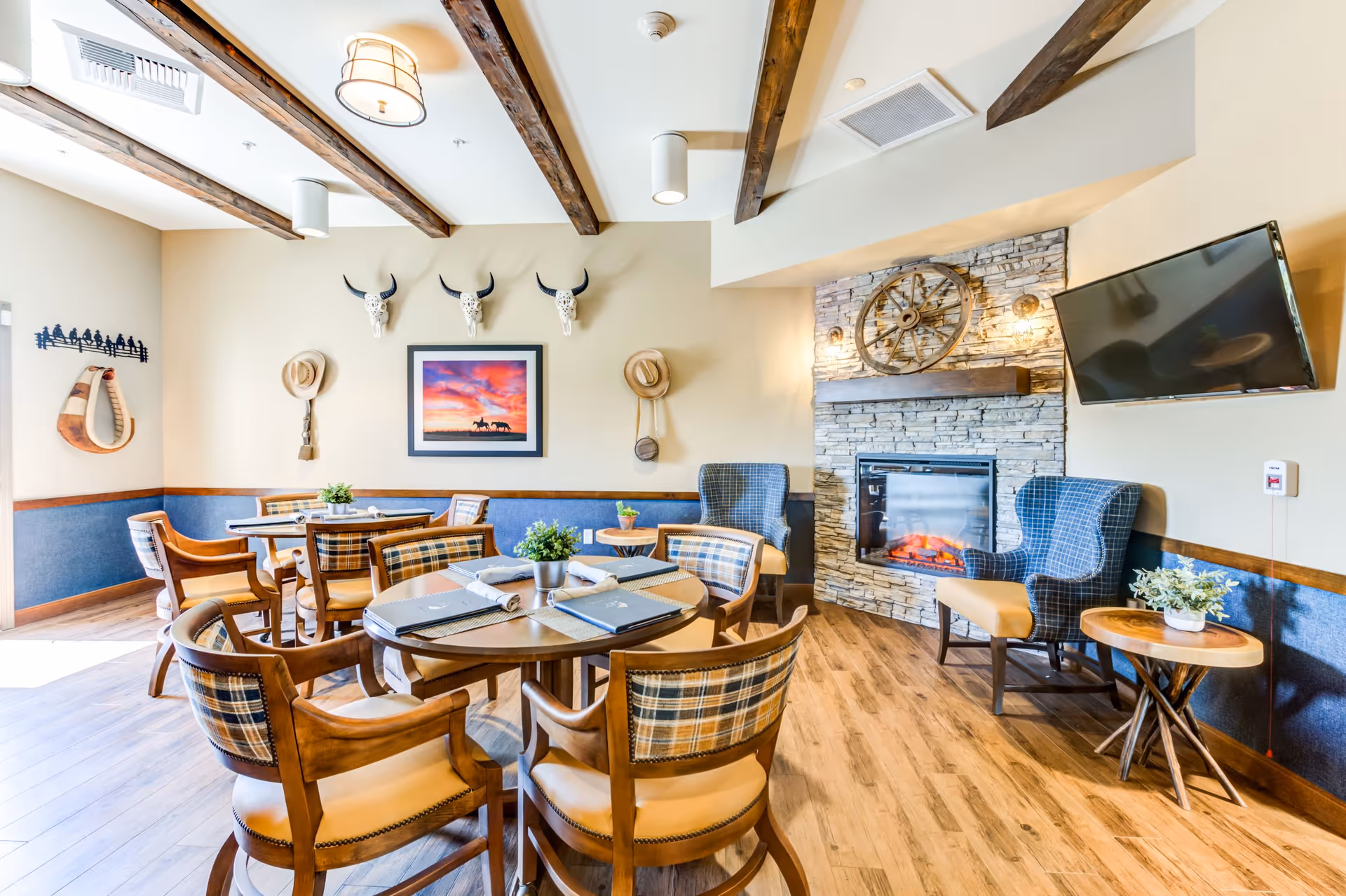 A cozy dining area with wooden tables and plaid cushioned chairs, a stone fireplace with a wagon wheel decoration above it, a wall-mounted TV, and rustic decor including hats and animal skulls on the wall.