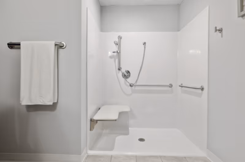 A clean, accessible shower area with white walls and a built-in fold-down seat. The shower has a handheld showerhead and two grab bars for safety. A white towel hangs on a metal towel rack mounted on the adjacent wall.