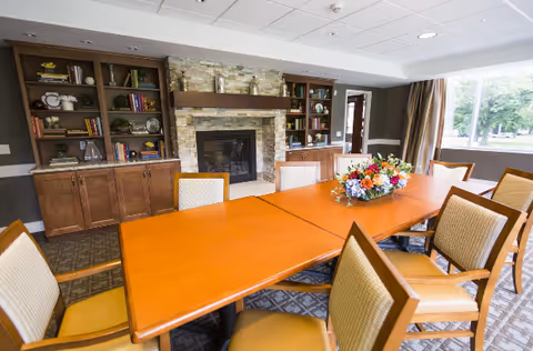 A well-lit room with a long wooden table surrounded by cushioned chairs. In the background, there is a stone fireplace flanked by built-in wooden bookshelves filled with books and decorative items. Large windows on the right side allow natural light to brighten the space.