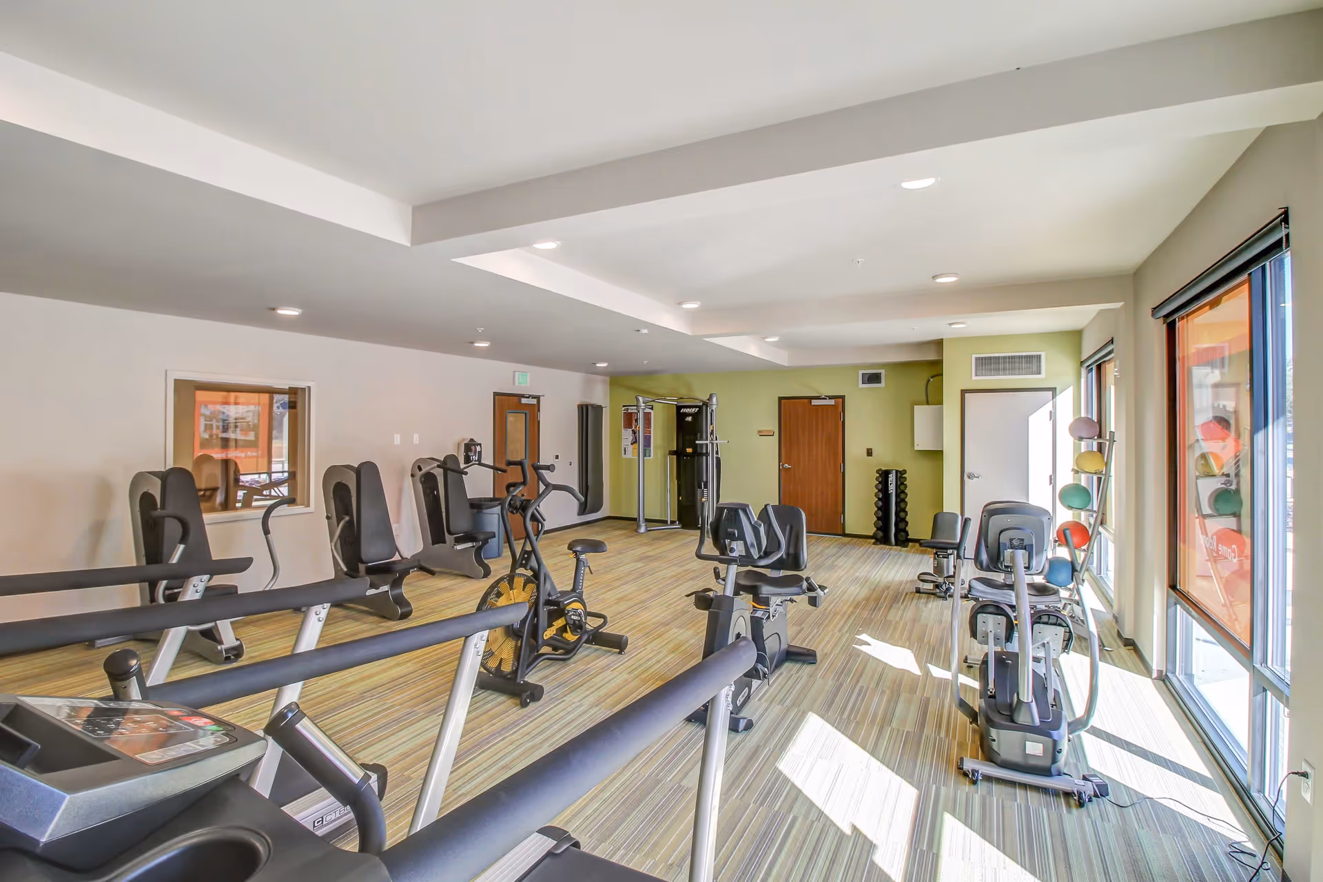 A bright fitness room with various exercise equipment including treadmills, stationary bikes, weight machines, and a rack of colorful medicine balls. The room has large windows allowing natural light to fill the space, light-colored walls, and a striped carpeted floor.