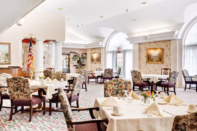A bright and elegant dining room with multiple tables covered in white tablecloths, each set with folded napkins, cups, and small floral centerpieces. The room features floral-patterned chairs, large windows with sheer curtains, framed paintings on the walls, and an American flag in the corner.