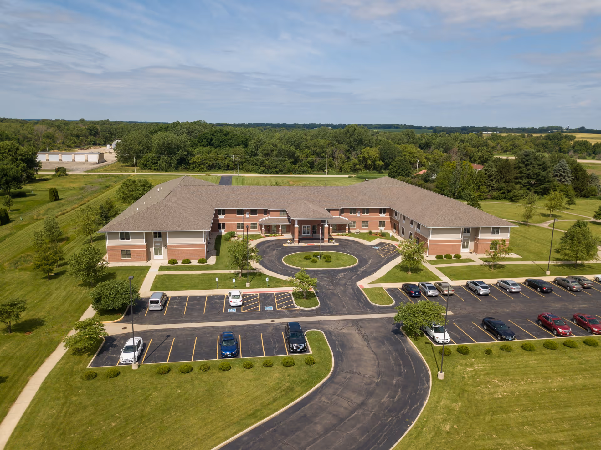 Aerial view of a two-story senior living facility building with a U-shaped design, surrounded by green lawns and trees. The building has a brown roof and brick exterior with white trim. There is a circular driveway with a small landscaped island in front of the main entrance, and parking lots with several cars parked. The surrounding area includes open fields and wooded areas under a partly cloudy sky.