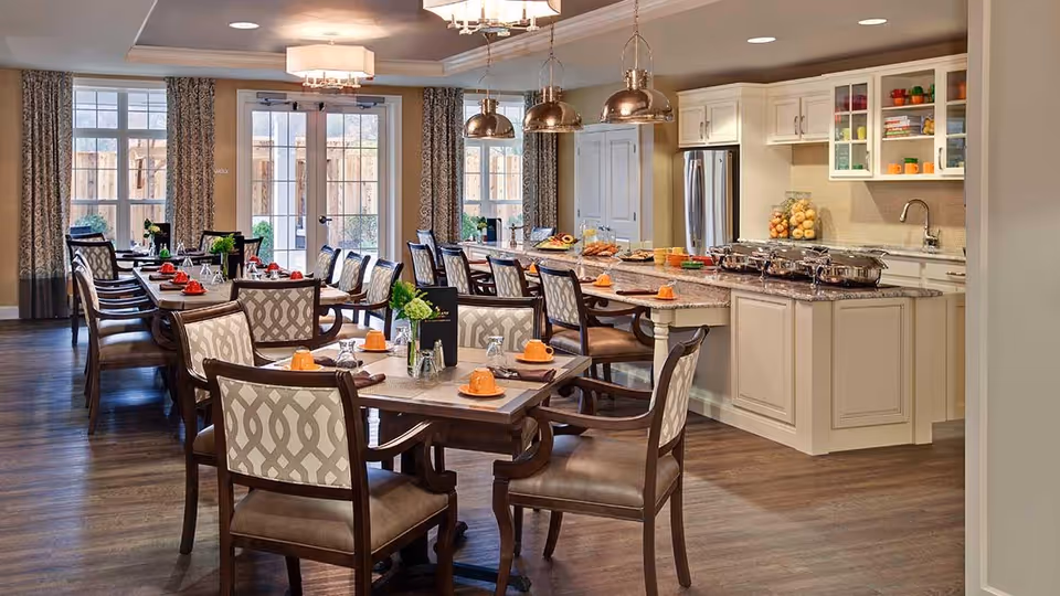 A spacious dining area with multiple wooden tables and chairs, each table set with orange cups, glasses, and napkins. The room features large windows with patterned curtains allowing natural light to enter. In the background, there is a kitchen area with white cabinetry, a granite countertop island, stainless steel refrigerator, and hanging pendant lights above the island. The floor is wooden, and the overall ambiance is warm and inviting.