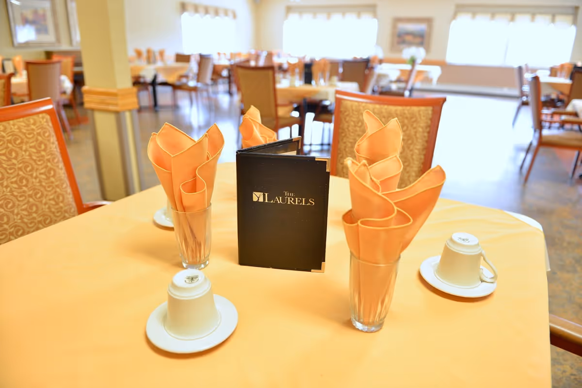A dining table set with folded orange napkins in clear glasses, upside-down white coffee cups on saucers, and a black menu with 'The Laurels' printed on it. The background shows a spacious dining room with multiple tables and chairs, softly lit by natural light from large windows.