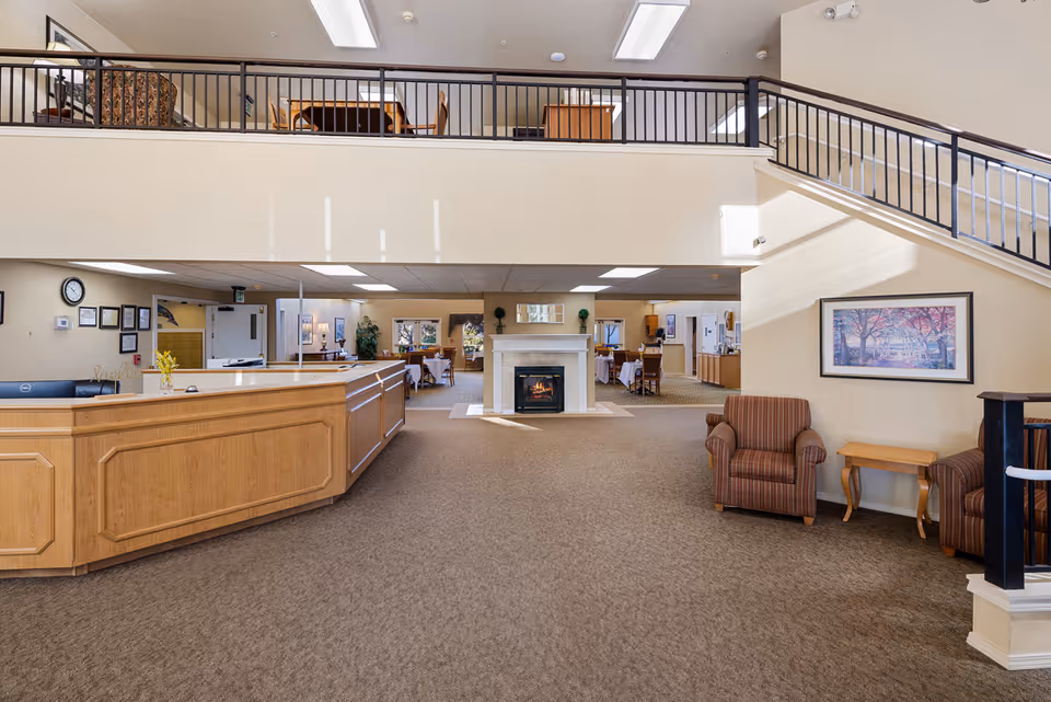 Interior view of a senior living facility lobby area with a wooden reception desk on the left, two striped armchairs and a small wooden table on the right, a fireplace in the center background, and a staircase with black railings leading to an upper floor with additional seating and tables.