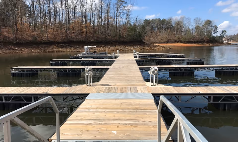 A wooden dock extending over a calm lake with multiple empty boat slips on either side. Leafless trees and some evergreen trees line the shore in the background under a partly cloudy blue sky.