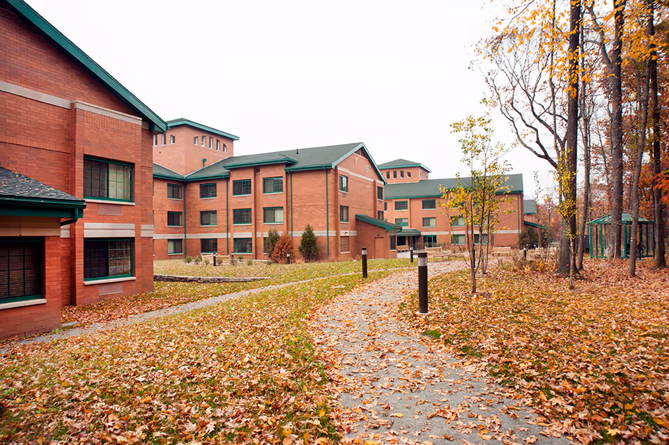 Brick multi-story senior living building with green roofs overlooking a leaf-covered courtyard and walking paths in autumn.