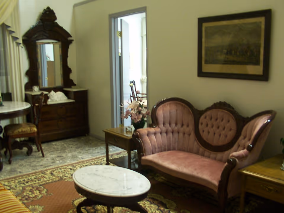 A vintage-style living room with a pink tufted velvet sofa, a round marble-top coffee table, a wooden side table with a flower vase, an ornate wooden dresser with a mirror, a round table with a chair, and a framed picture on the wall.