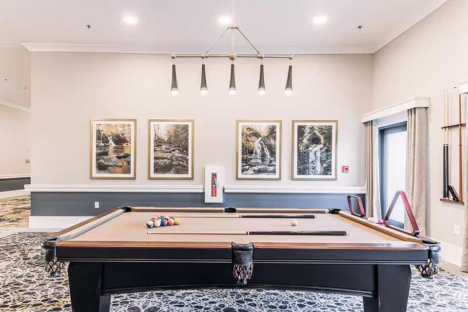 A well-lit room featuring a pool table with billiard balls and cues arranged on it. The room has a patterned carpet and neutral-colored walls adorned with four framed nature-themed pictures. A modern light fixture with five hanging lights is suspended above the pool table. There is a window with curtains on the right side and a fire extinguisher mounted on the wall.