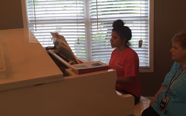 A young woman in a red shirt is playing a white piano in a room with large windows covered by white blinds. An older woman wearing a blue top with a lanyard is sitting nearby, watching her play.