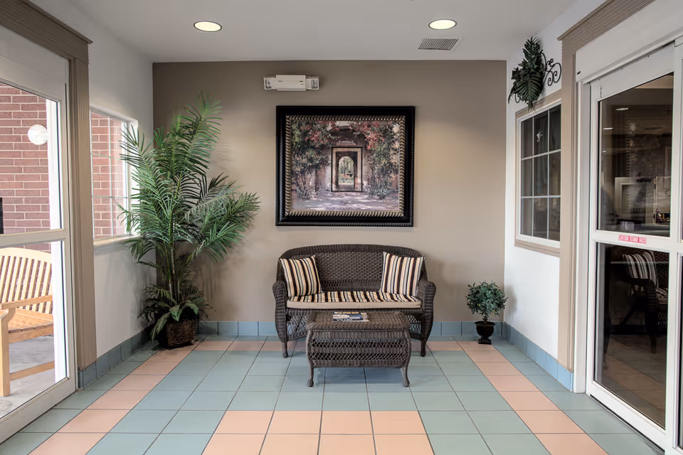 A small seating area with a wicker loveseat and matching coffee table, both with striped cushions, placed against a beige wall. Above the loveseat hangs a framed painting depicting an archway with greenery. There are two potted plants on either side of the loveseat, and large windows and glass doors surround the area, allowing natural light to enter.