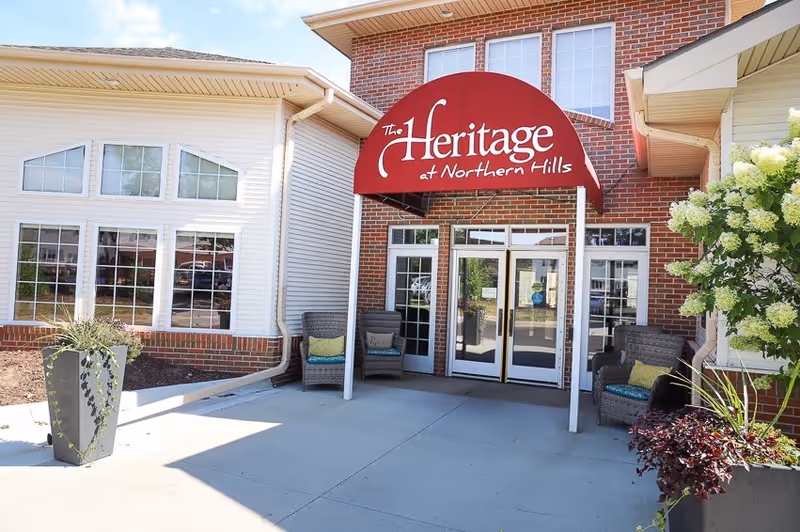 Entrance of The Heritage at Northern Hills facility with a red awning displaying the facility name. The entrance has double glass doors, two wicker chairs with cushions on either side, and plants near the doorway. The building exterior is a combination of brick and siding.