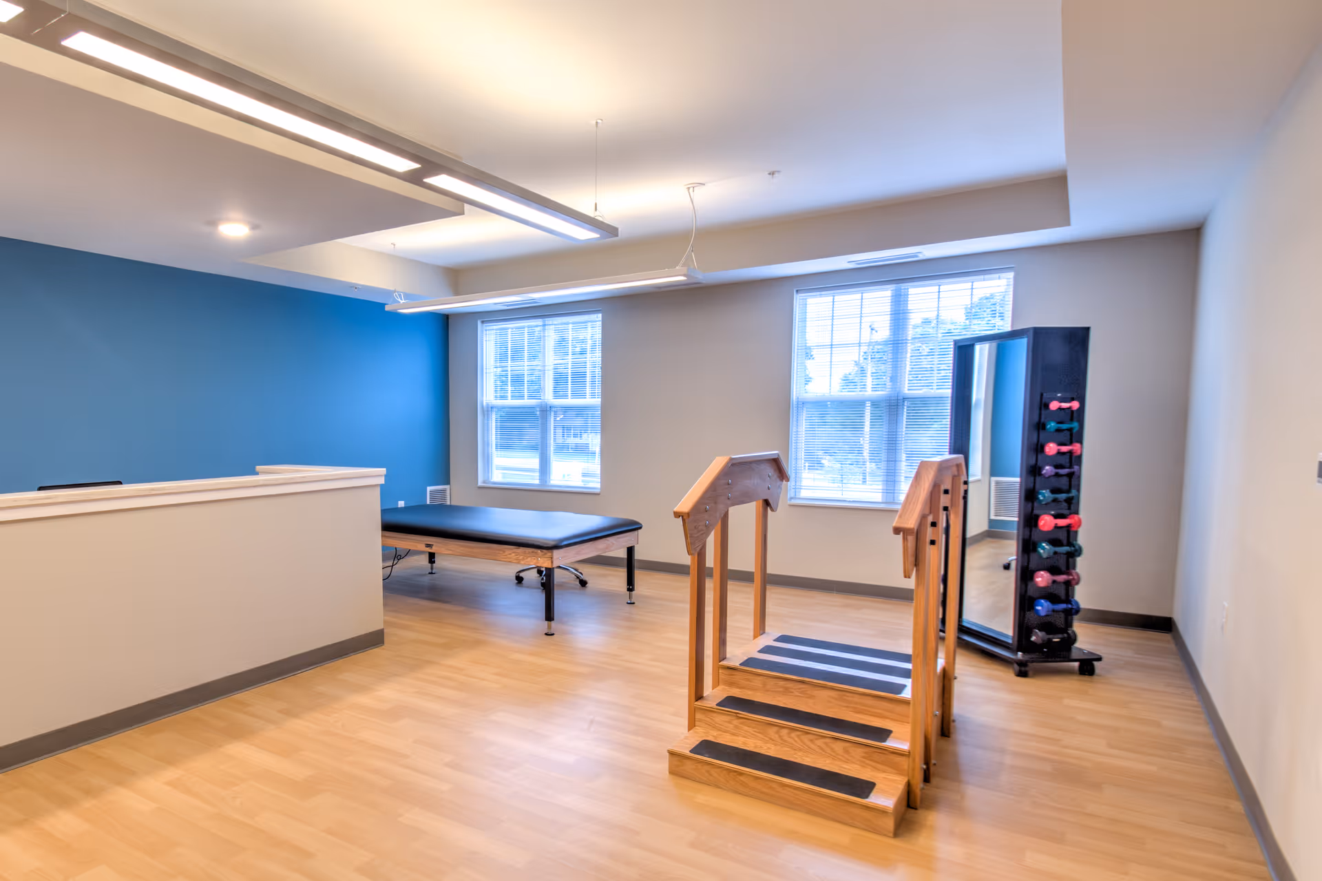 A bright room with light wood flooring and white walls, featuring a blue accent wall. The room contains a black padded therapy table, a wooden set of stairs with handrails for physical therapy, and a rack with colorful dumbbells next to a full-length mirror. Two large windows allow natural light to fill the space.