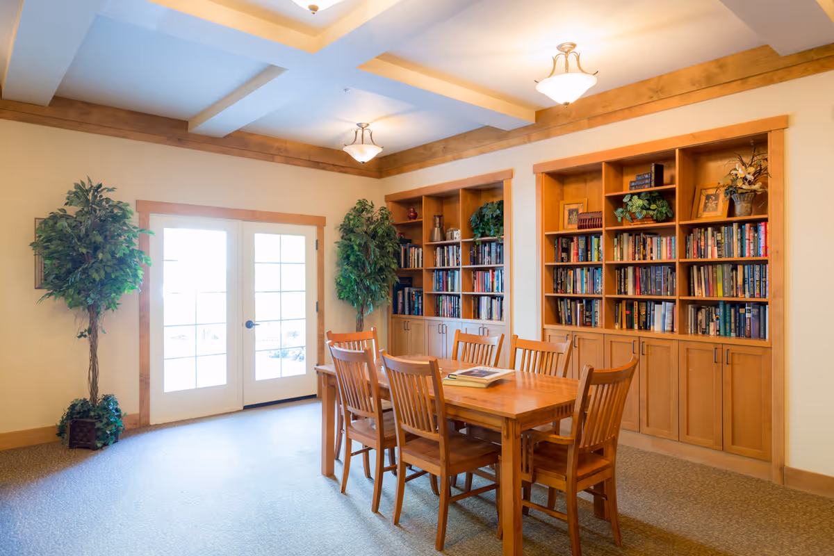 A well-lit room with a wooden table and six matching wooden chairs. Behind the table are two large built-in wooden bookshelves filled with books and decorative plants. There are two tall potted plants on either side of a set of double glass doors with wooden frames. The ceiling has recessed lighting and wooden trim accents.