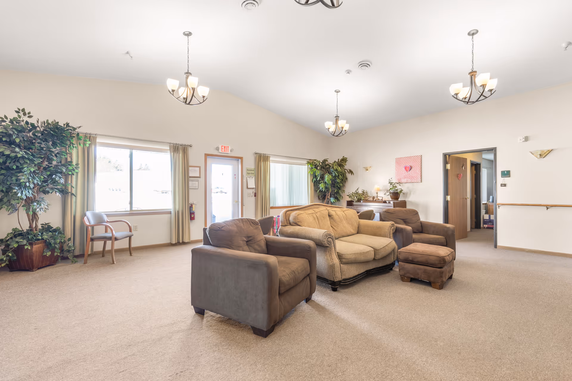 Bright common living room with sofas, armchairs, potted plants, and chandeliers in a senior living facility.