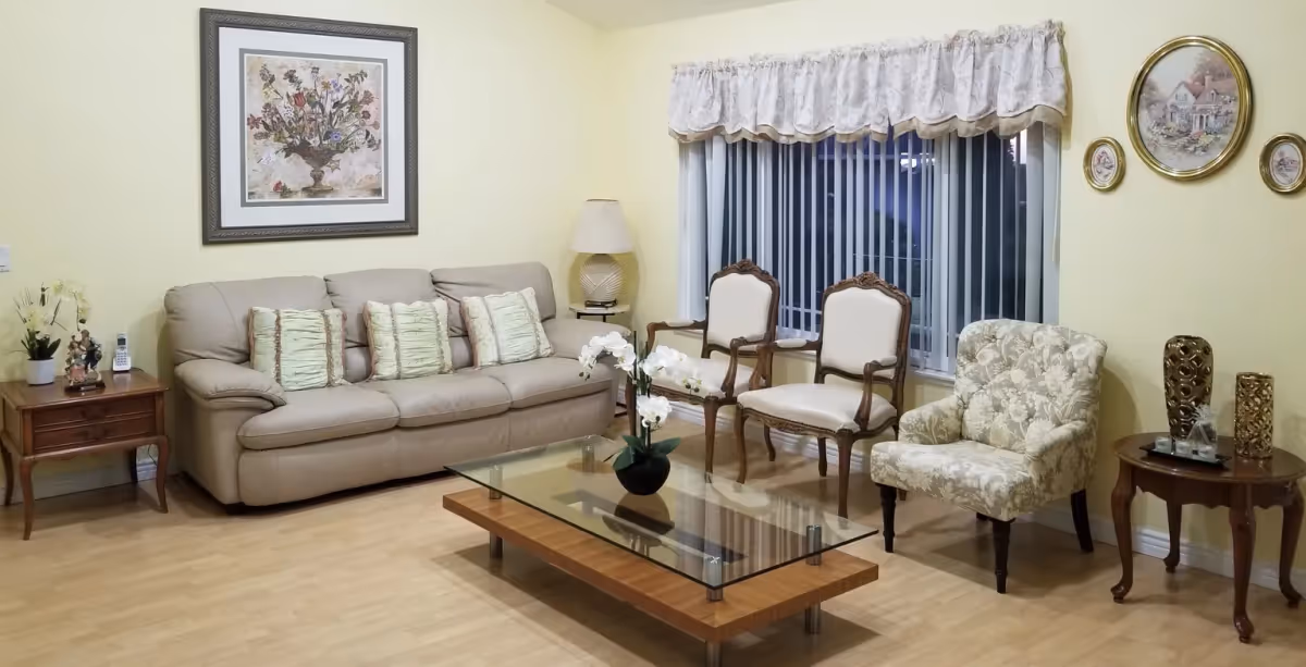A cozy living room with a beige leather sofa adorned with four light green pillows, two classic wooden armchairs with white cushions, and a floral-patterned upholstered armchair. A glass coffee table with a wooden base holds a small white orchid plant. The room features light yellow walls decorated with framed floral and house-themed artwork, a window with vertical blinds and a white valance, and two wooden side tables with decorative items and a lamp.