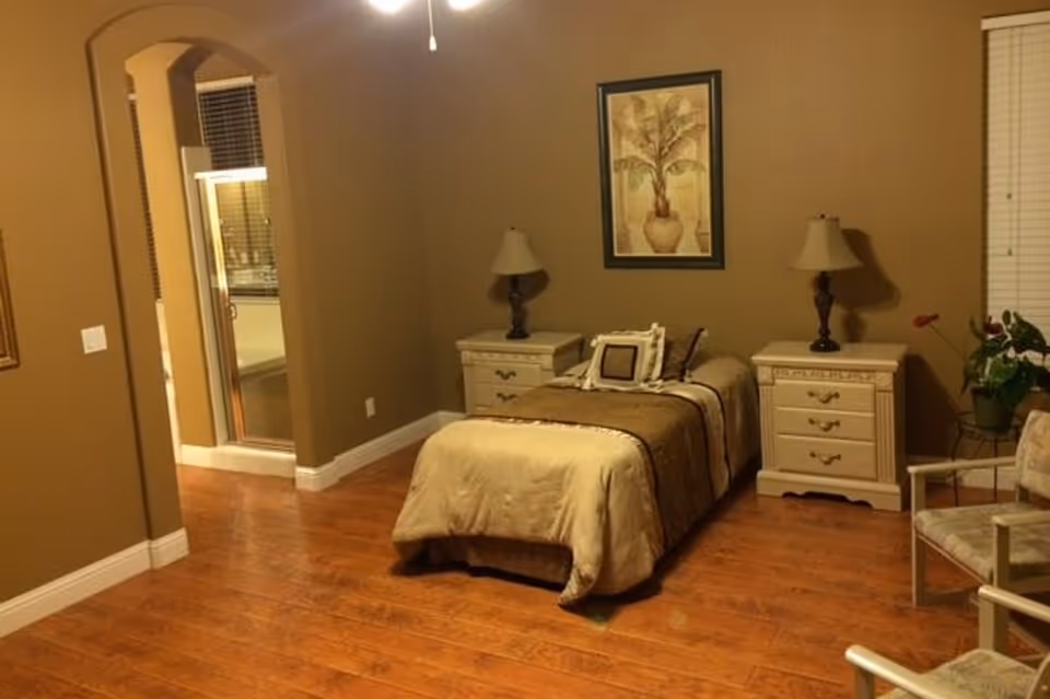 A cozy bedroom with a single bed covered in beige and brown bedding, flanked by two white nightstands each with a lamp. There is a framed picture of a plant on the wall above the bed. The room has wooden flooring, a window with blinds, two chairs, and a small plant on a stand. An open doorway leads to a bathroom with a glass shower enclosure.