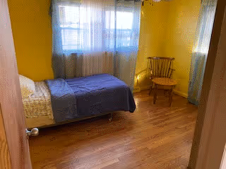 A small bedroom with yellow walls and wooden flooring. The room contains a single bed with a white pillow and a blue blanket, positioned against the left wall under a window with sheer white curtains. There is a wooden chair with armrests placed near the corner on the right side of the room.