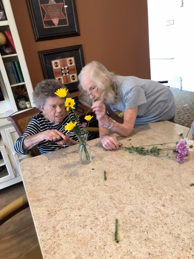 Two elderly women arranging yellow and purple flowers in a small glass vase on a beige table in a room with brown walls and framed artwork.