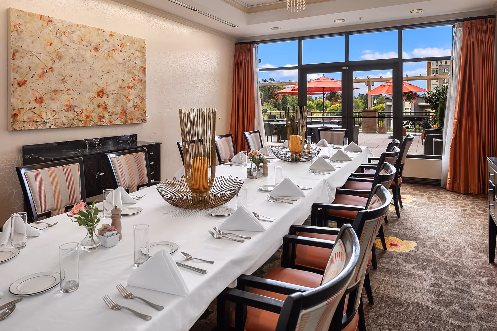 A formal dining room with a long table covered with a white tablecloth, set with folded white napkins, plates, glasses, and silverware. The room has upholstered chairs around the table, a large abstract painting on the wall, and large windows with orange curtains that open to an outdoor patio with red umbrellas and greenery.