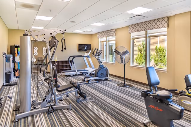 A well-lit fitness room with exercise equipment including a multi-station weight machine, two treadmills, an exercise bike, and a standing fan. The room has large windows with valances, striped carpet flooring, and beige walls.