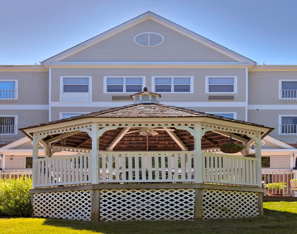 A white wooden gazebo with a shingled roof and ceiling fan, situated on a grassy lawn in front of a multi-story beige building with multiple windows and balconies under a clear blue sky.