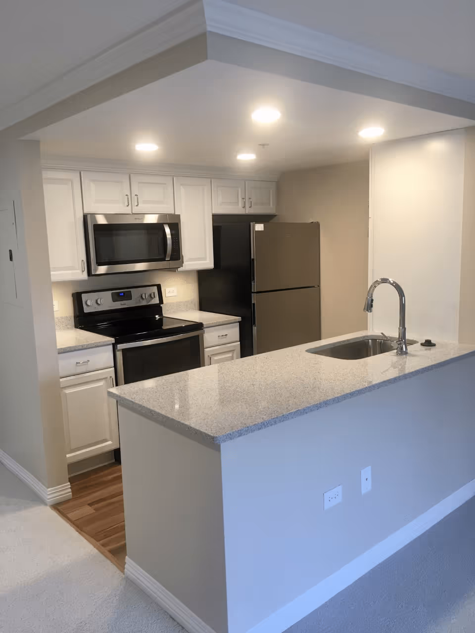 Modern kitchen area with white cabinets, stainless steel microwave, stove, and refrigerator. A light-colored granite countertop with an integrated sink and chrome faucet extends into a breakfast bar. The kitchen is illuminated by recessed ceiling lights and has beige walls and a mix of carpet and wood flooring.