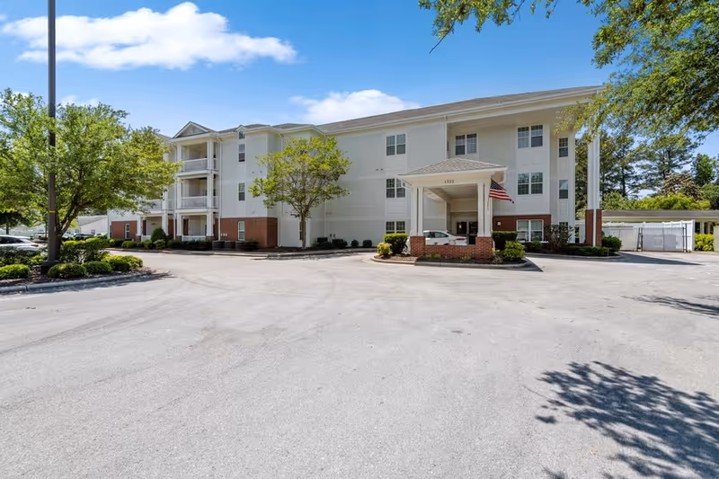 Exterior view of a three-story senior living facility building with white siding and brick accents. The building has a covered entrance with an American flag, surrounded by trees and a paved driveway under a blue sky with some clouds.