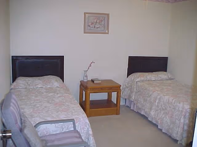 A simple bedroom with two twin beds separated by a wooden nightstand. Each bed has a floral bedspread and a dark headboard. On the nightstand, there is a small vase with a single flower and a book. A framed picture hangs on the wall above the nightstand. A chair with a striped cushion is partially visible in the foreground.