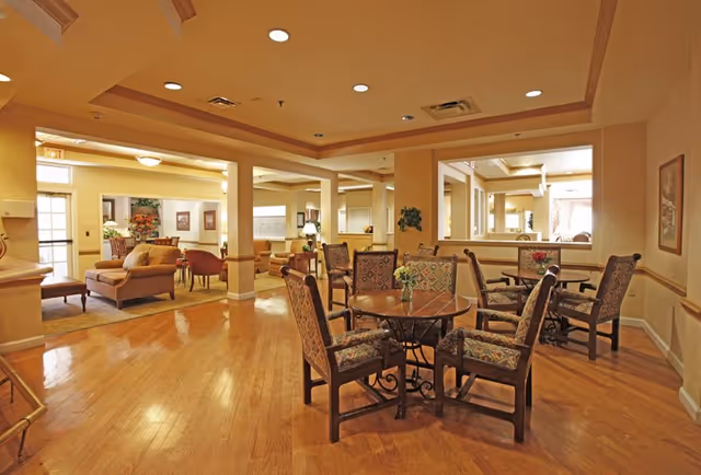 A spacious and warmly lit common area in a senior living facility featuring wooden floors, round tables with upholstered chairs, and a seating area with sofas and armchairs. The walls are painted in soft beige tones with decorative molding, and recessed ceiling lights provide illumination. There are large windows and mirrors enhancing the openness of the space.