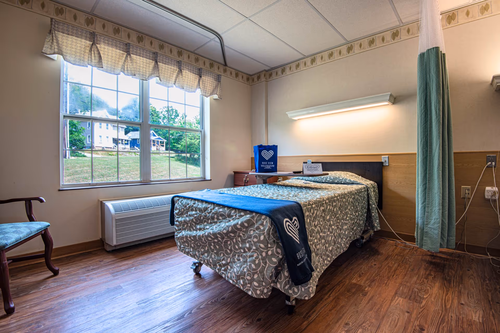 A single bed in a nursing facility room with patterned bedding and a blue blanket featuring the Rose View logo. The room has a large window with a view of green grass and houses outside, a wooden chair with blue upholstery, a wall-mounted light above the bed, and a privacy curtain on the right side. The floor is wooden, and there is an air conditioning unit below the window.