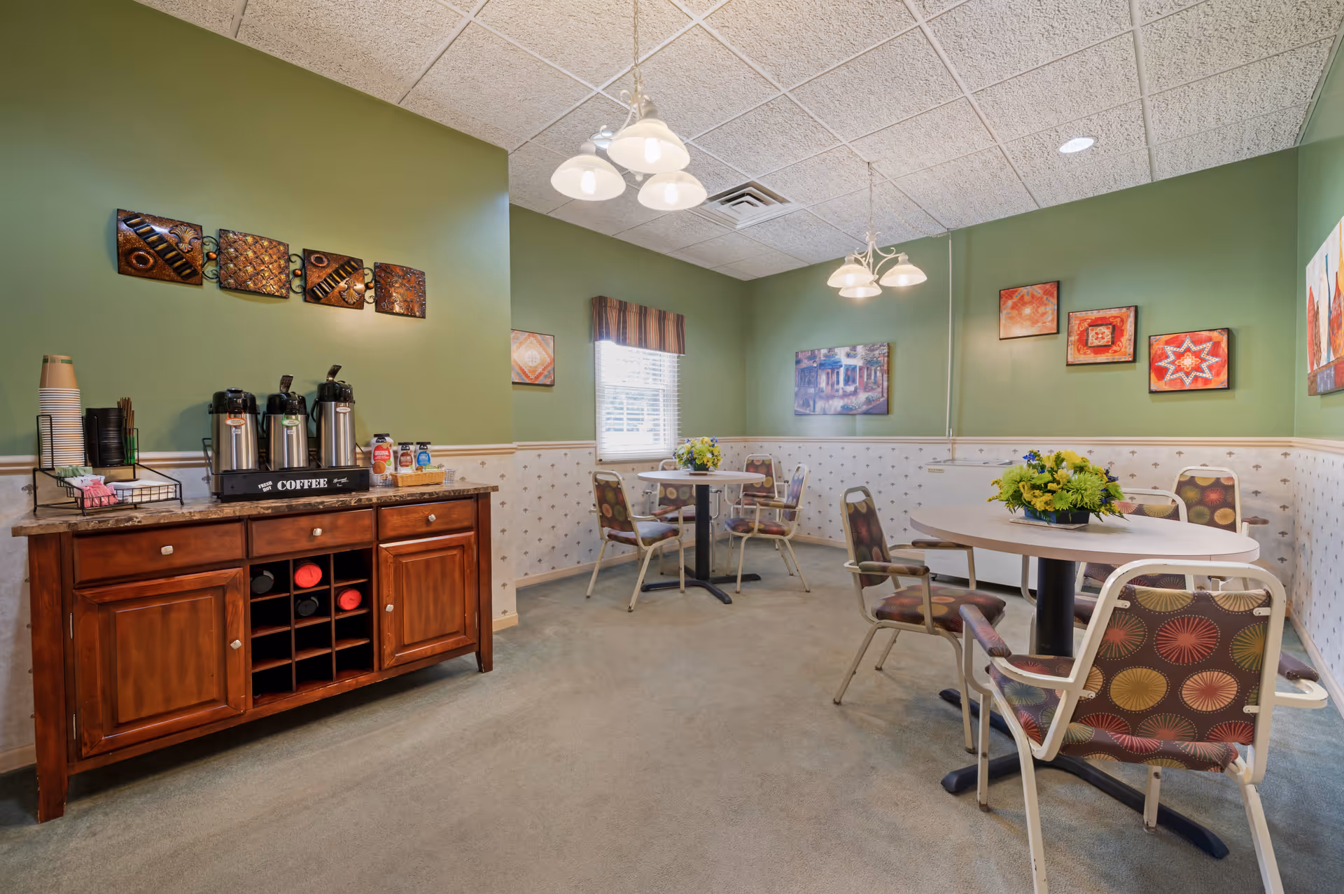 A cozy dining area in an assisted living community with green walls and patterned wallpaper. The room features round tables with chairs that have colorful circular patterns on the upholstery. A wooden sideboard holds coffee dispensers, cups, and condiments. The walls are decorated with various framed artworks, and two ceiling light fixtures illuminate the space.