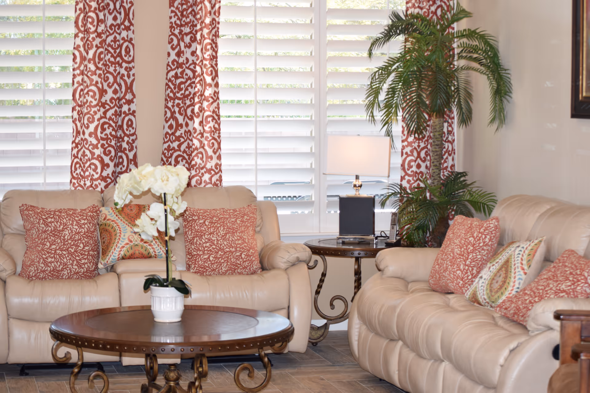 A cozy living room with two beige leather sofas adorned with red and patterned throw pillows. A round wooden coffee table with a white potted orchid is in the center. Behind the sofas are windows with white plantation shutters and red patterned curtains. A side table with a lamp and a cordless phone sits between the sofas, and a tall green potted plant is in the corner.