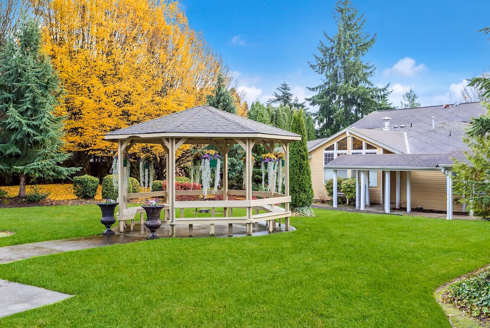 A wooden gazebo with hanging flower baskets on a green lawn in front of a beige building and autumn trees.