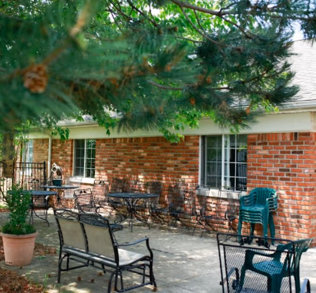 Outdoor patio area with metal tables and chairs, a bench, and stacked green plastic chairs next to a brick building. There are trees providing shade over the seating area.