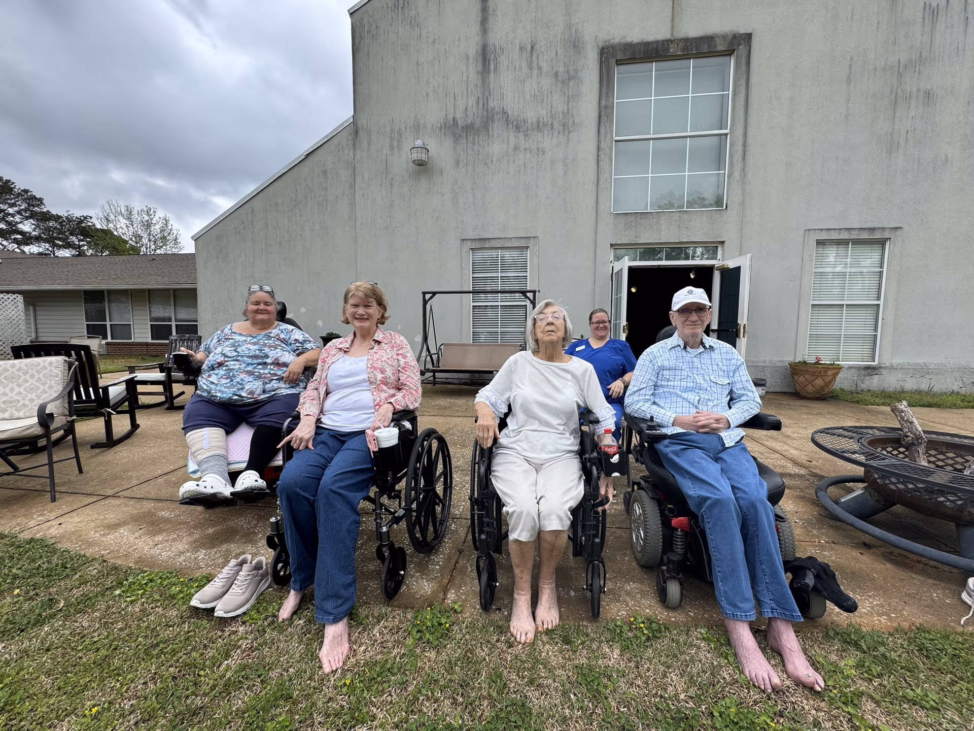 Four elderly individuals sitting outside on a concrete patio in front of a building, three in wheelchairs and one in a motorized wheelchair. A caregiver in blue scrubs is standing behind them. The sky is cloudy and there is grass in the foreground.