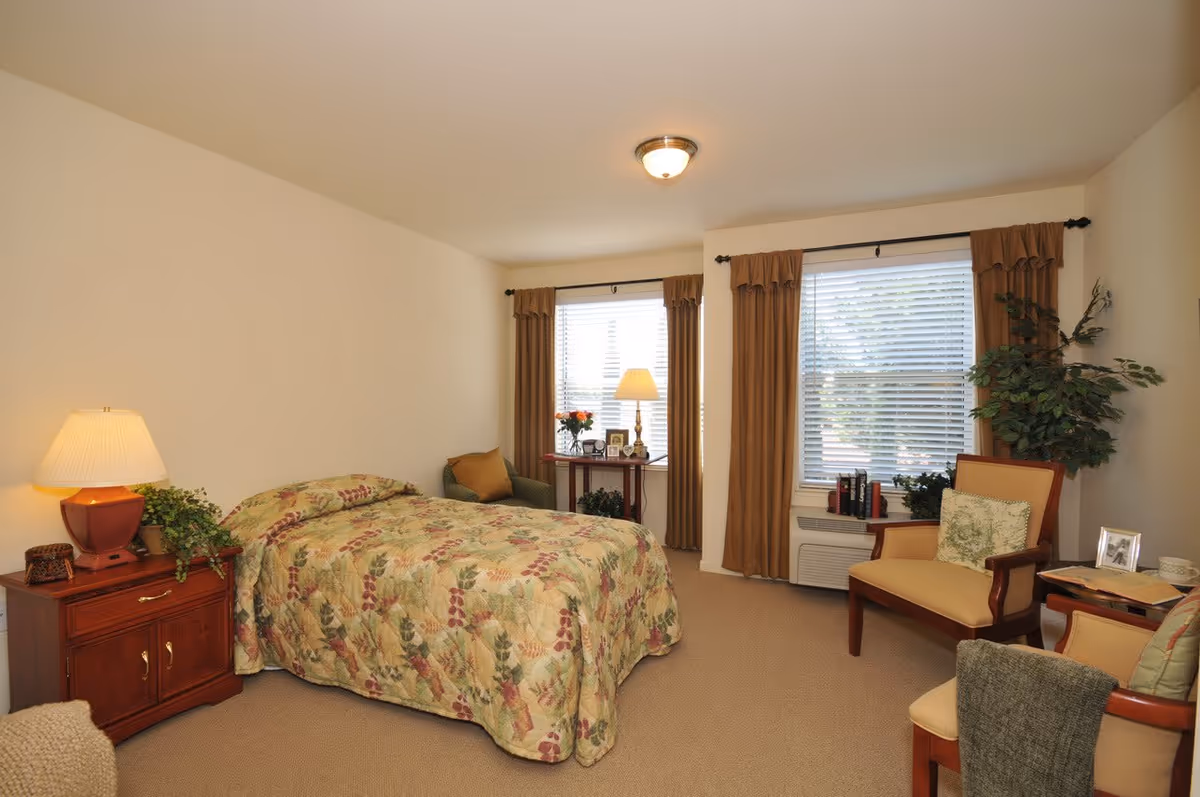 A cozy bedroom with a bed covered in a floral patterned bedspread, a wooden nightstand with a lamp and plant, two large windows with brown curtains, a small table with a lamp and picture frames, and two armchairs with cushions and a small side table.