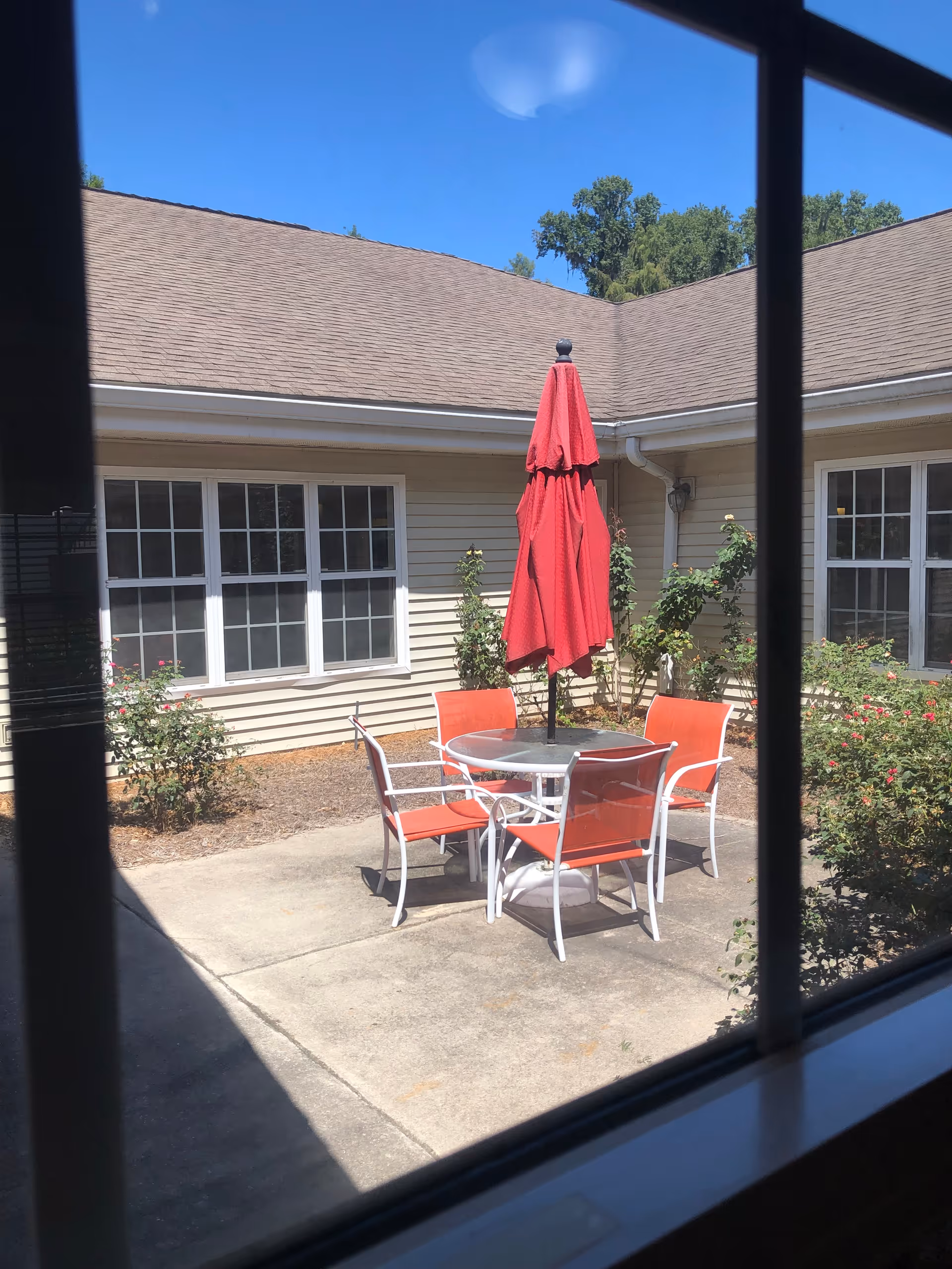 View through a window of a small outdoor patio area with a round glass table, four orange chairs, and a closed red umbrella. The patio is surrounded by beige building walls with windows and some green bushes and plants under a clear blue sky.