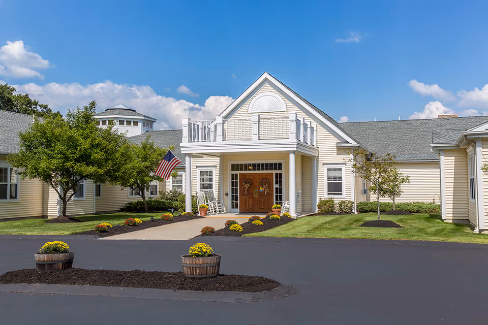 Front exterior view of a single-story senior living facility building with beige siding, a peaked roof, and a covered entrance with white columns. There are two rocking chairs on the porch, an American flag on a pole, and landscaped flower beds with yellow and red flowers. The sky is clear with some clouds.