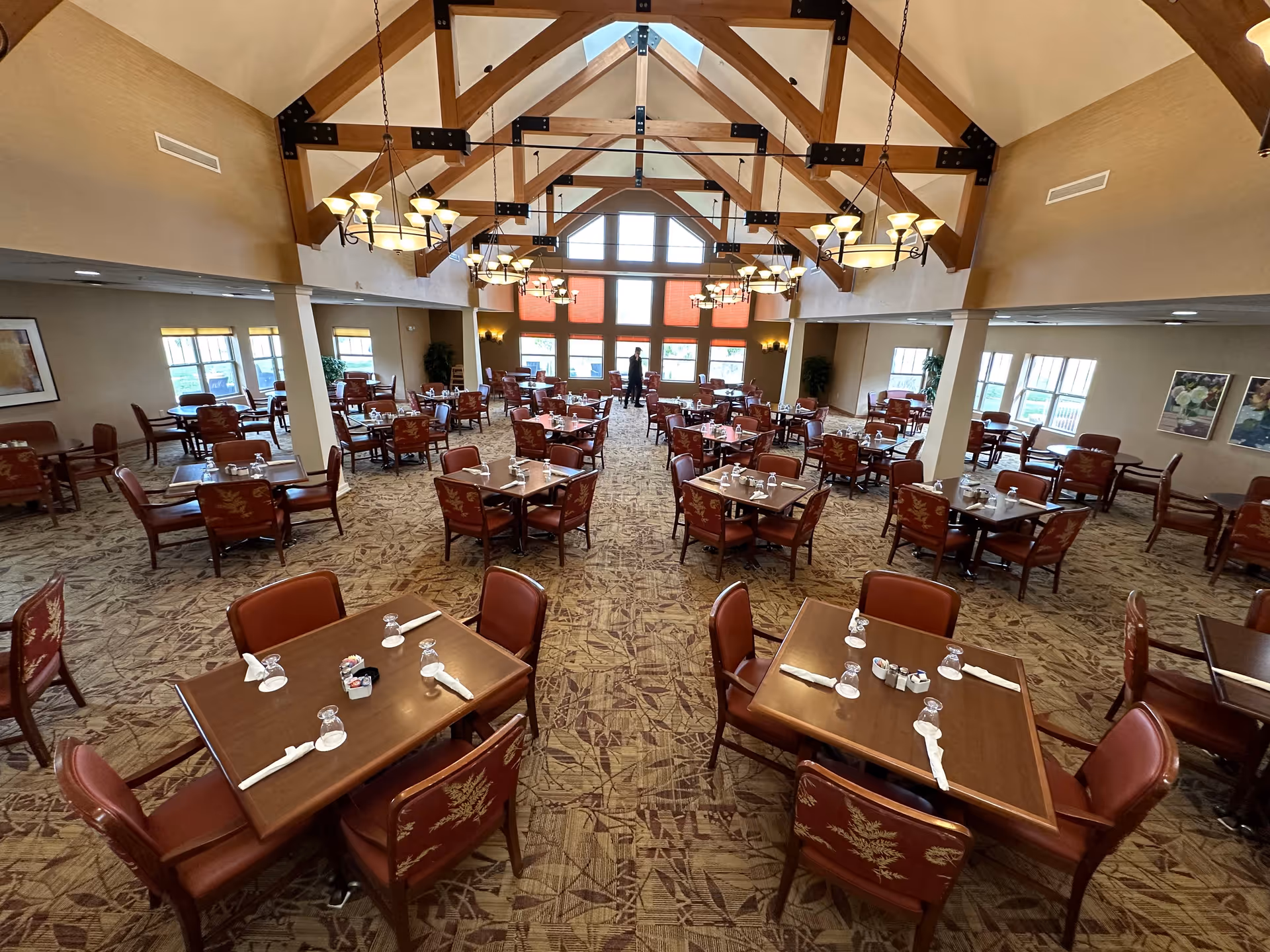 Large, well-lit dining room with multiple set tables and chairs, decorative chandeliers, and exposed wooden ceiling beams.