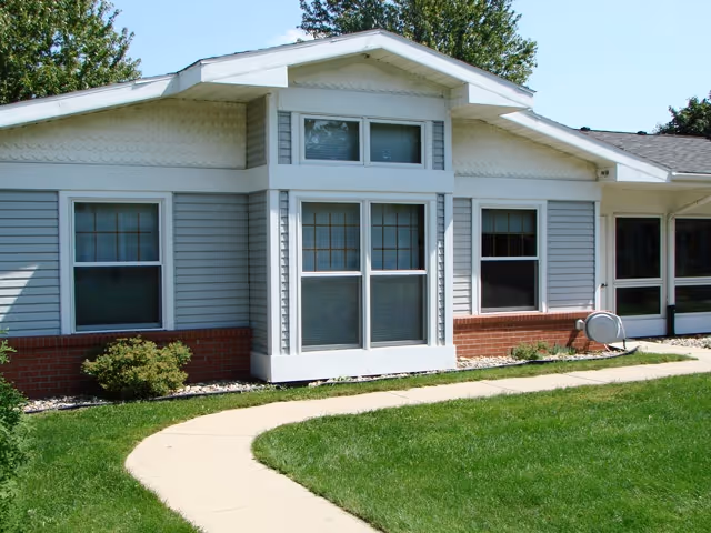 Exterior view of a single-story building with gray siding and red brick at the base. The building has multiple windows and a small curved concrete walkway leading to the entrance. There is green grass and some bushes in front of the building under a clear blue sky.