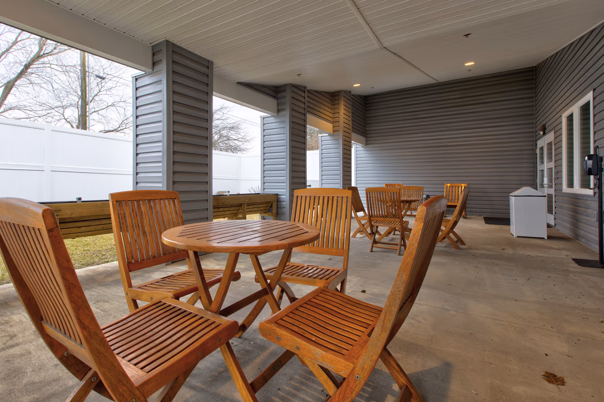 Covered outdoor patio area with multiple wooden tables and chairs arranged on a concrete floor. The space is enclosed on one side by gray siding walls and on the other by white fencing, with some trees visible outside the fence.