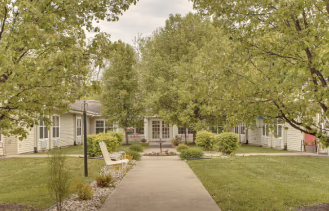 A paved walkway leads through a courtyard with green grass, trees, and bushes on either side. There are benches along the path and beige buildings with white doors and windows surrounding the courtyard under a cloudy sky.